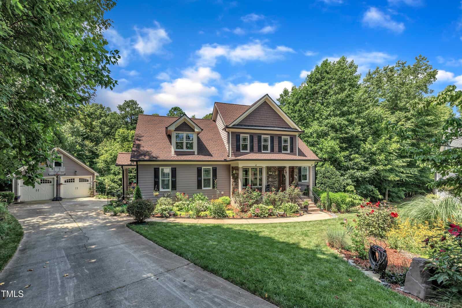 A two-story house with brown siding and a red roof sits amid green trees and a landscaped yard. A curved driveway leads to a detached garage. The lawn and garden beds are neatly maintained under a partly cloudy sky.