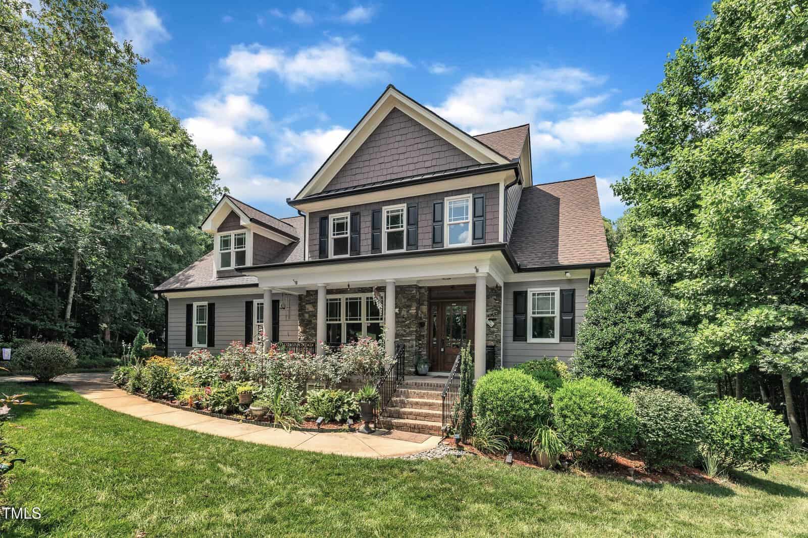 Two-story house with gray siding, white trim, and dark shutters. Covered front porch with columns, brick steps, landscaped front yard with bushes and flowers, and surrounded by trees under a blue sky with clouds.