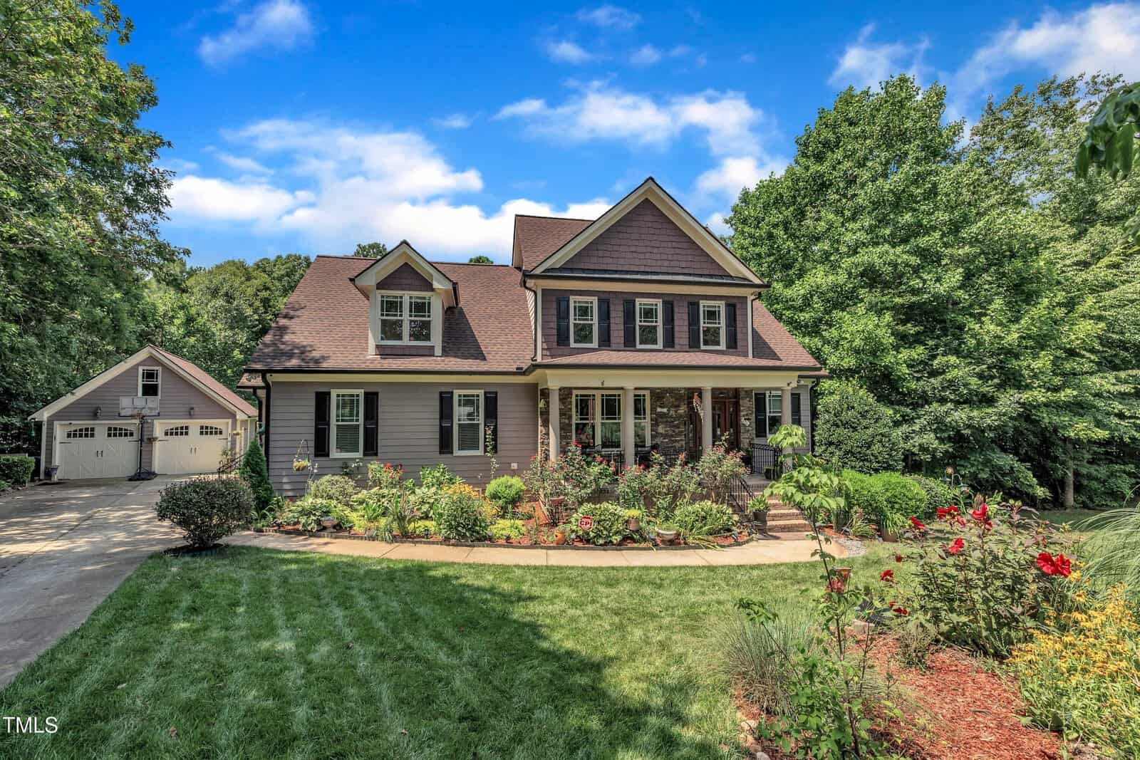 A two-story house with a covered front porch, gabled roof, and dormer windows is surrounded by a well-kept lawn and landscaped garden. A detached garage sits to the left, and trees border the property.