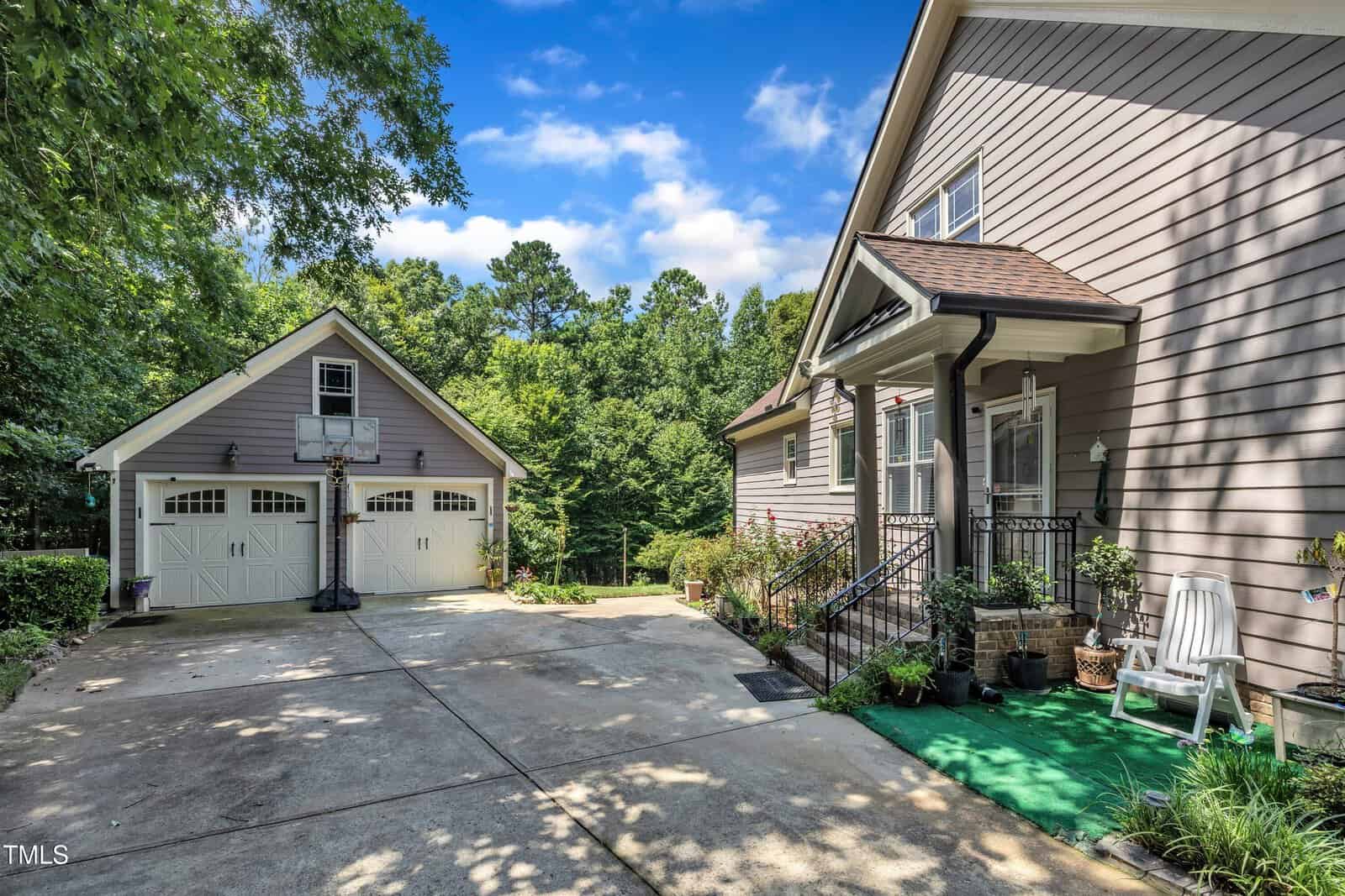 A driveway leads to a two-car garage with white doors and a basketball hoop. The house has beige siding, a small porch with potted plants, and a white chair on a green outdoor rug. Trees fill the background under a blue sky.