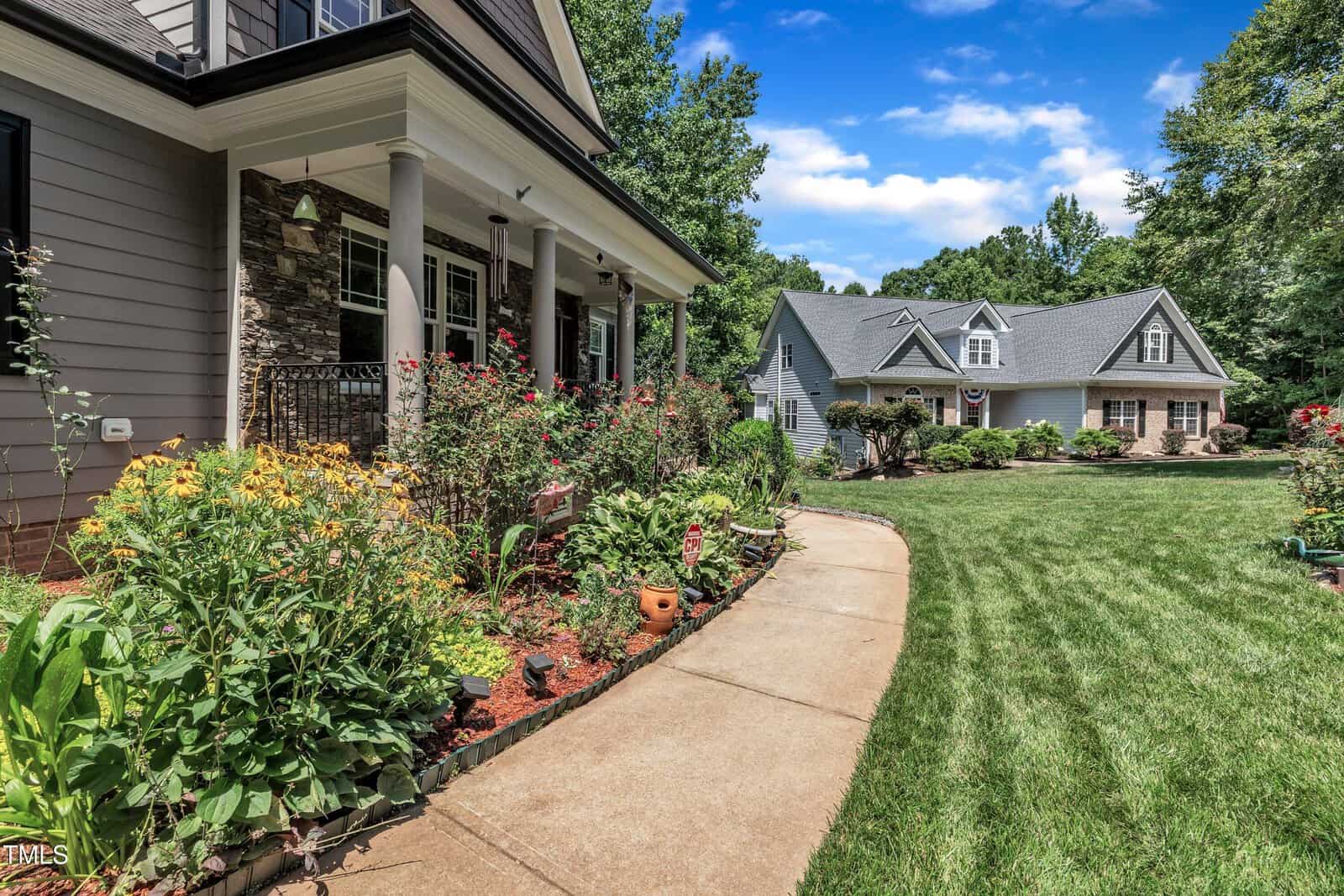 A curved sidewalk leads past colorful flower beds to a large house with a covered porch; another gray house sits in the background, surrounded by green grass and trees under a blue sky.