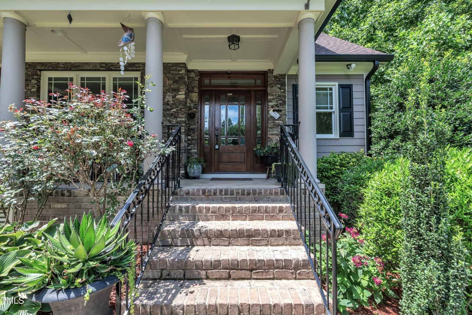 Front steps with wrought-iron railings lead to a wooden door, surrounded by flowers, shrubs, and stone accents on a house’s porch.