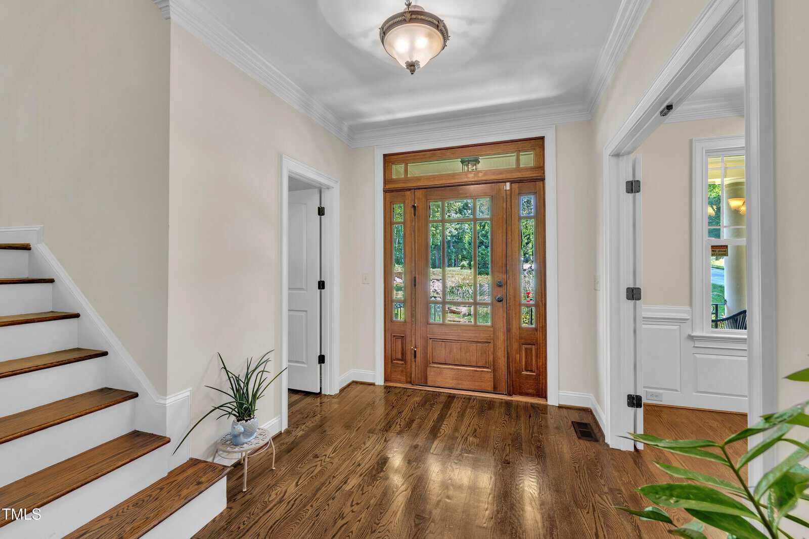 A bright foyer with wood floors, a wooden front door with glass panels, white walls, a staircase with wooden steps, and green plants on the floor and in the corner.