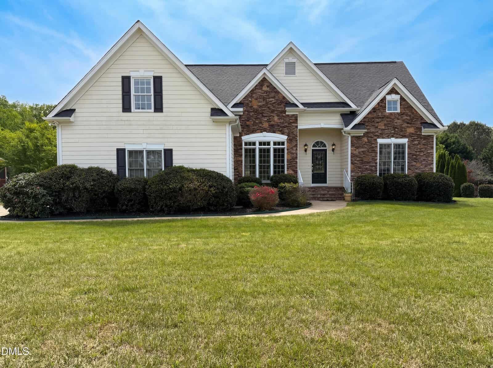 A single-family house with stone and white siding exterior, black shutters, and a dark front door, surrounded by shrubs and a large green lawn under a blue sky.