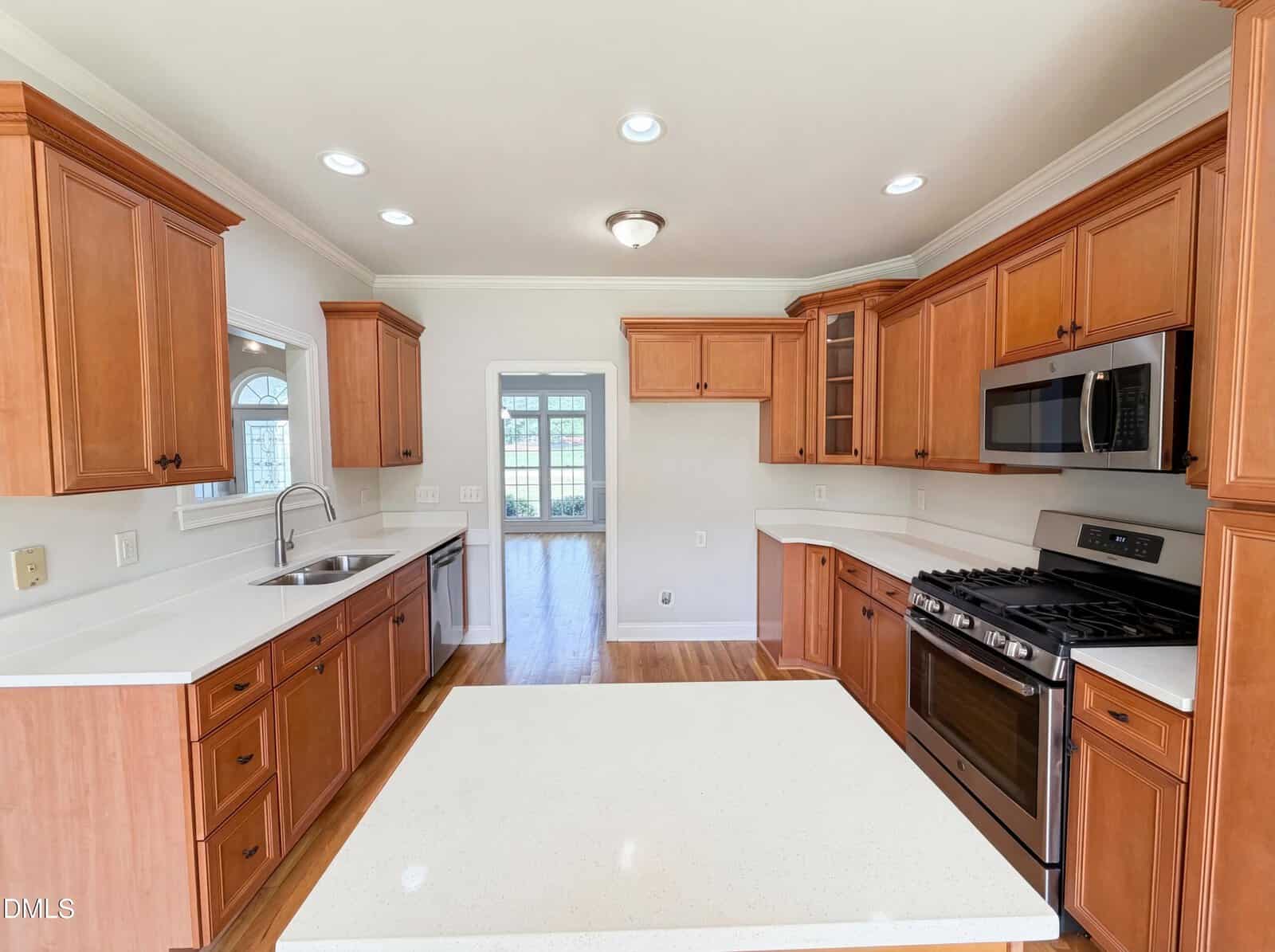 Modern kitchen with wooden cabinets, white countertops, stainless steel appliances including a microwave and gas stove, a central island, and a window above the sink. An open doorway leads to a bright adjoining room.
