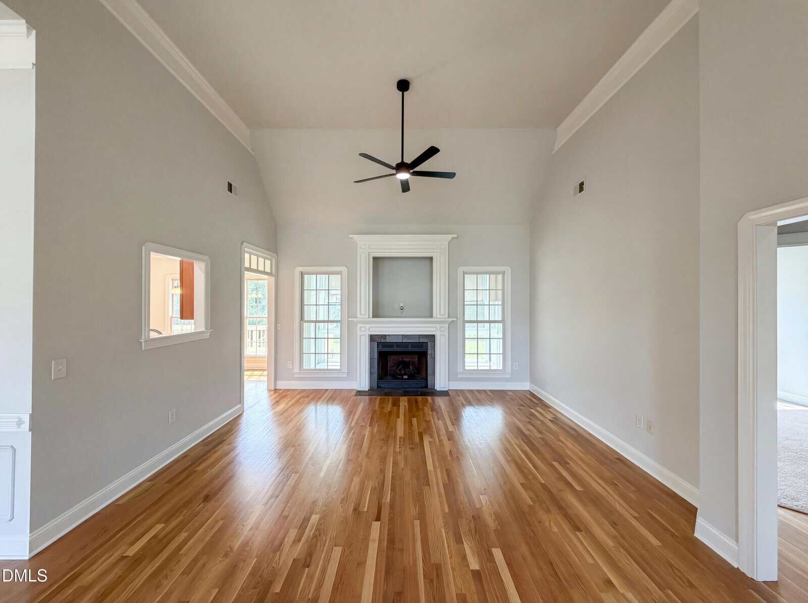 A spacious living room with light hardwood floors, high vaulted ceilings, a black ceiling fan, white walls, and a white fireplace centered between two tall windows. Natural light enters from multiple windows.