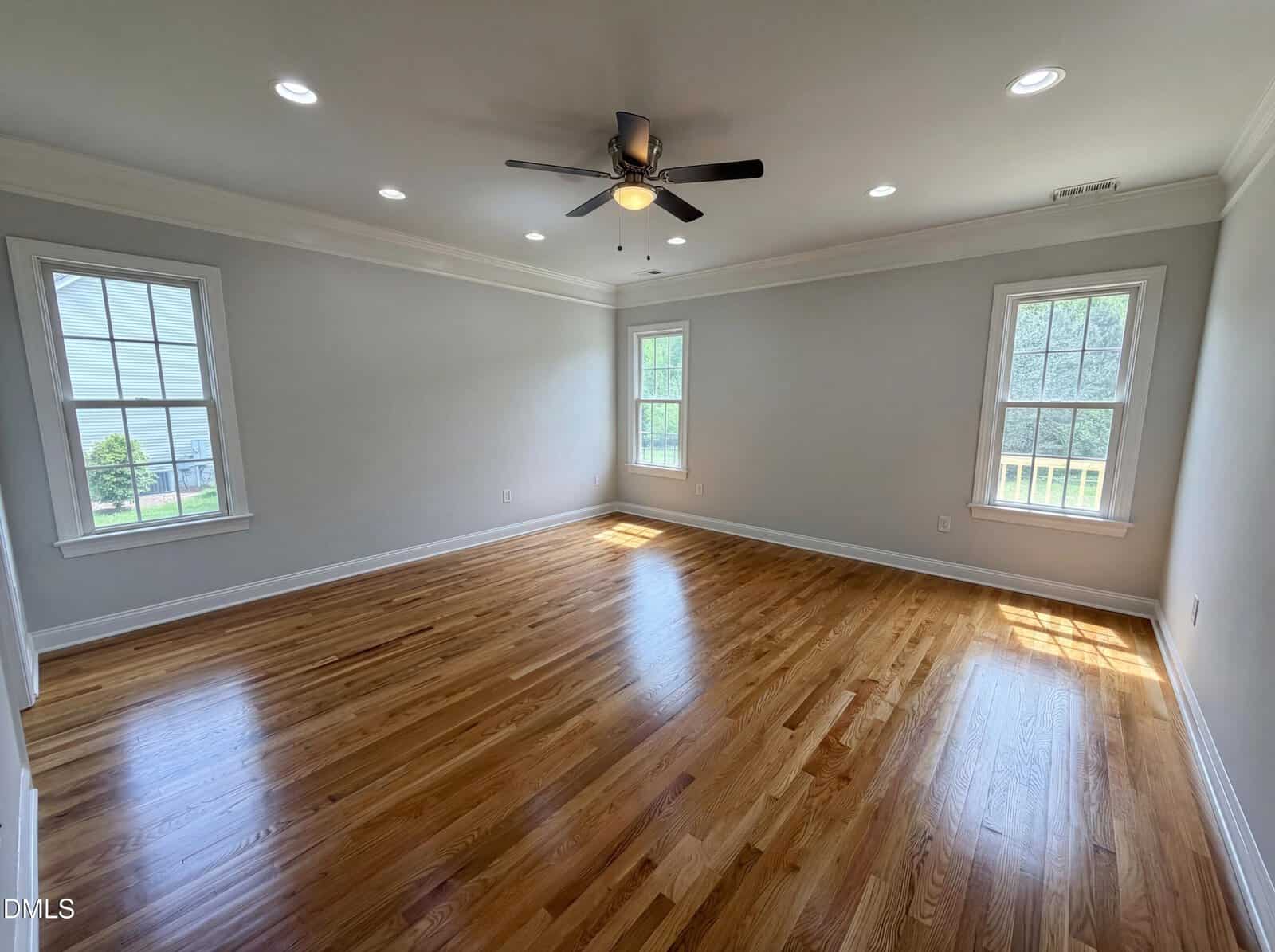 Empty room with light-colored walls, hardwood floors, four windows letting in natural light, a ceiling fan with lights, and recessed ceiling lights. The room is clean, spacious, and unfurnished.
