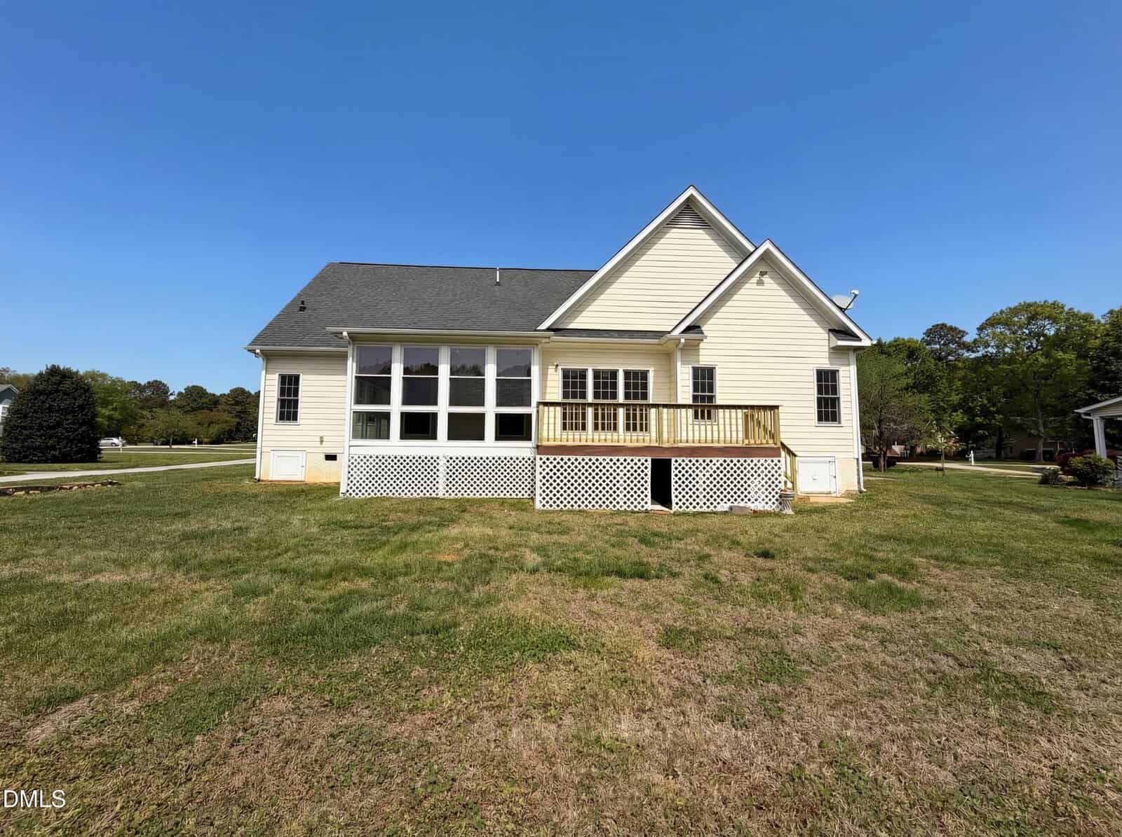 A single-story white house with a dark roof, a screened porch, and a small wooden deck, set on a large grassy lawn under a clear blue sky.