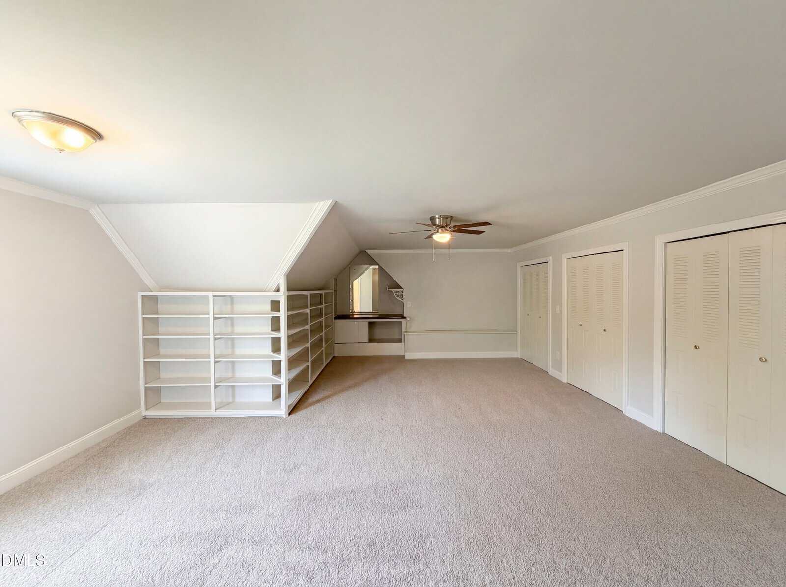 Spacious carpeted room with white walls, built-in shelves on the left, a ceiling fan, two closet doors on the right, and a small nook with a mirror at the back.
