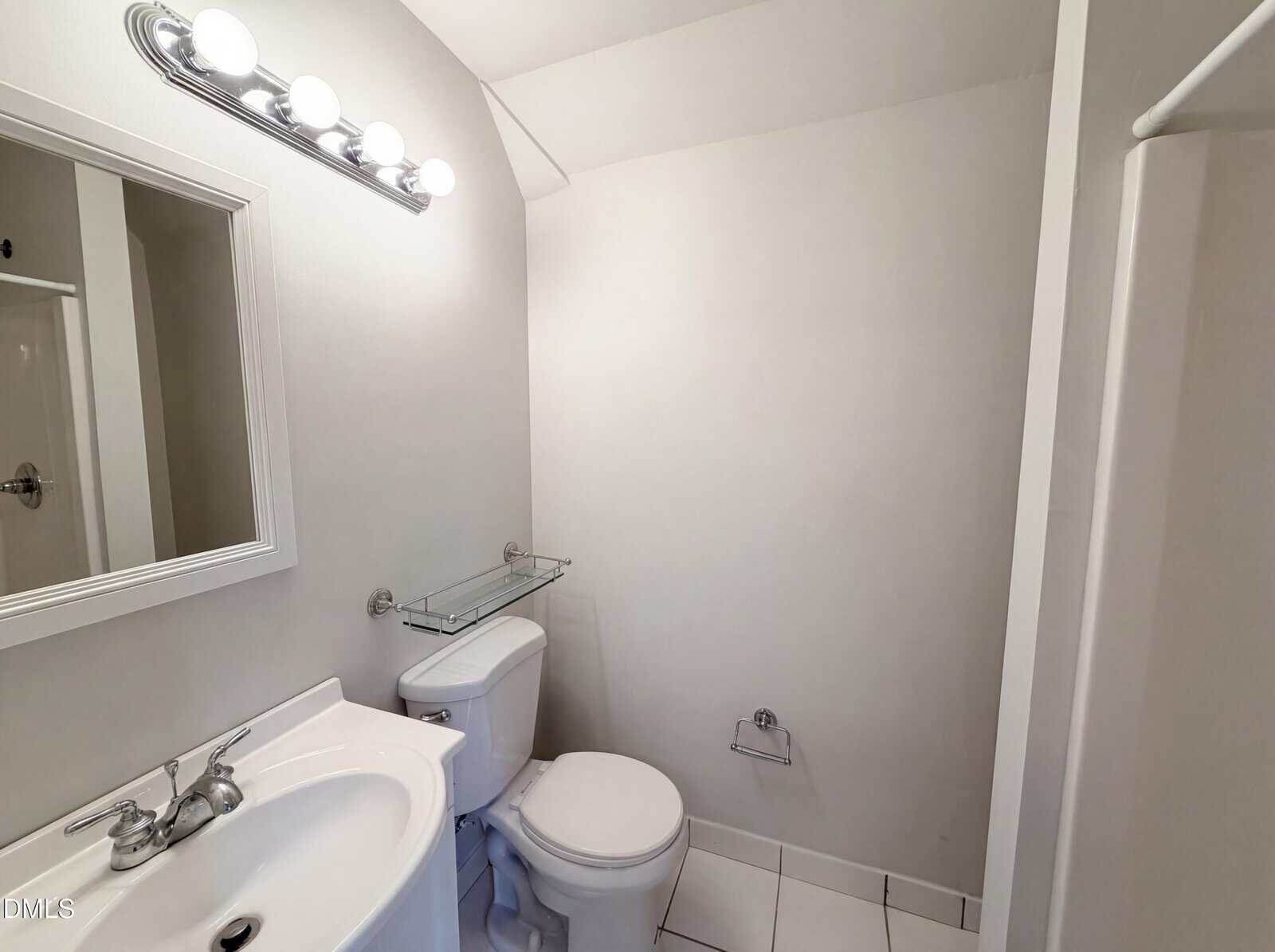 A small, white bathroom with a toilet, sink, mirror, and wall-mounted light fixture. A towel holder is on the wall next to the toilet, and a glass shelf is above the toilet.