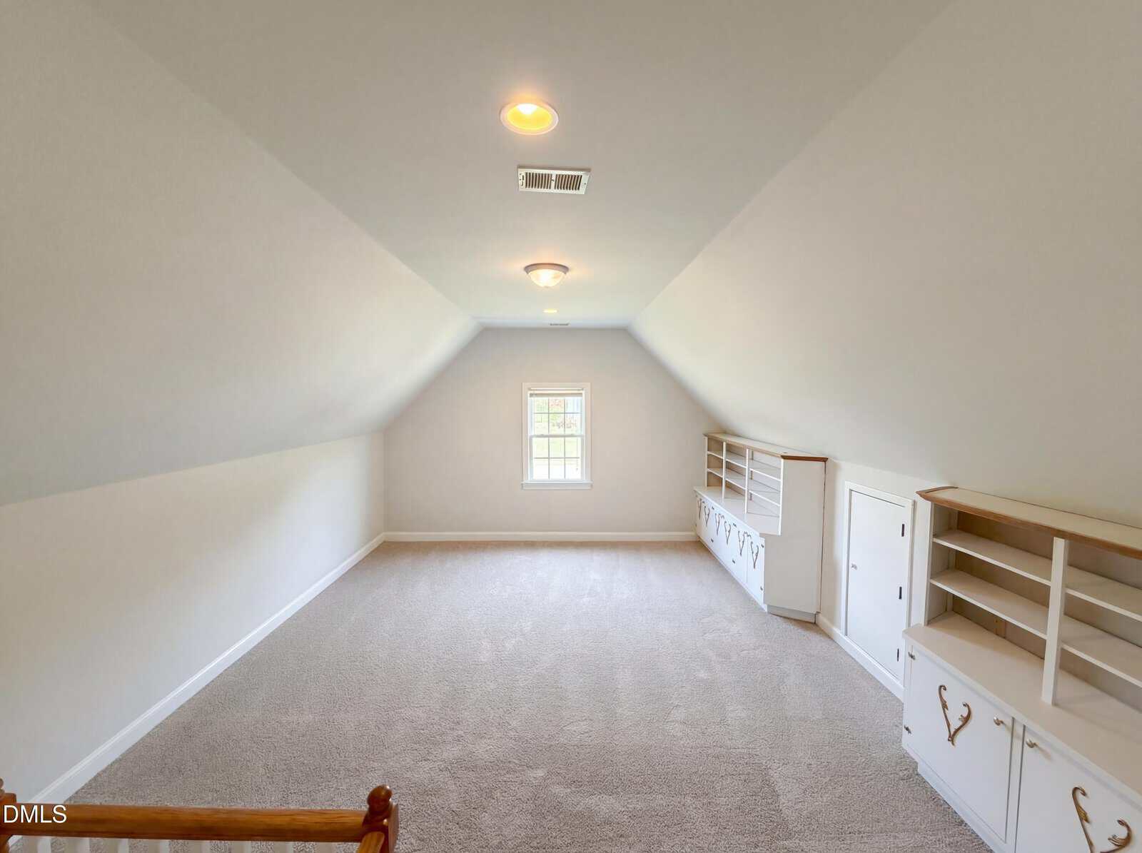 Bright attic room with light carpet, sloped walls, a small window at the end, built-in white shelves and cabinets on the right side, and recessed ceiling lights. The space is empty and well-lit.