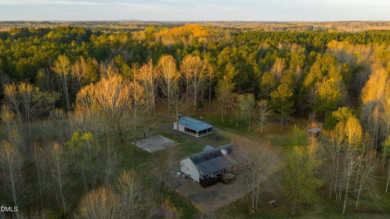 Aerial view of a small house and an outbuilding surrounded by leafless and evergreen trees, with a cleared yard area, in a rural forested landscape at sunset.