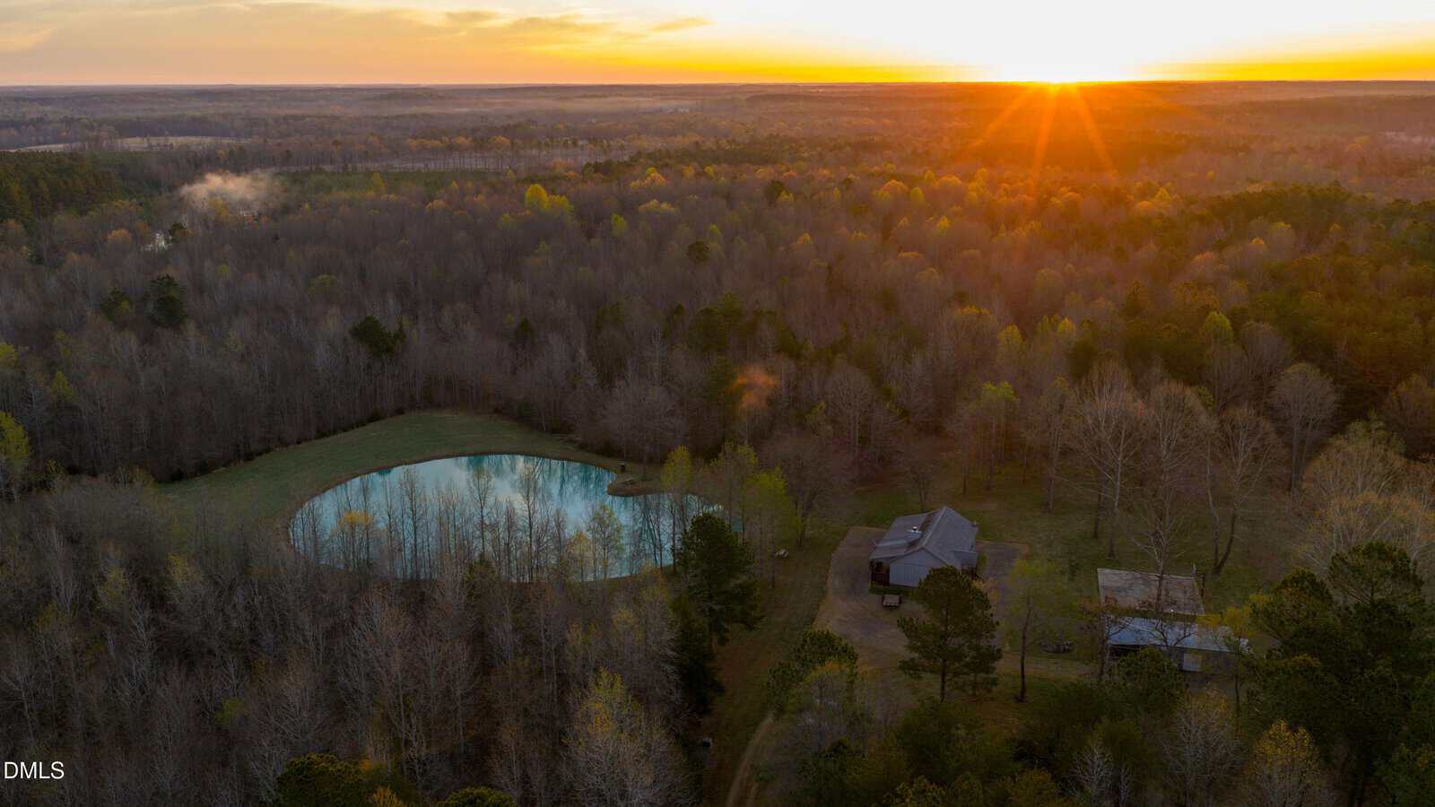 Aerial view of a house and pond surrounded by trees at sunset, with sunlight streaming over the forest and a clear sky above.