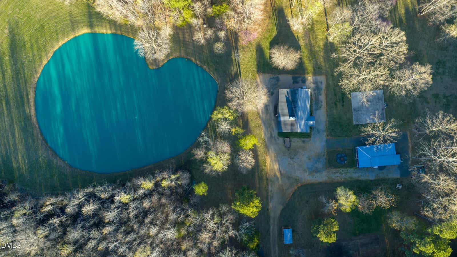 Aerial view of a rural property with a blue pond, a house with a gray roof, a small building, trees, and grassy areas under sunlight.