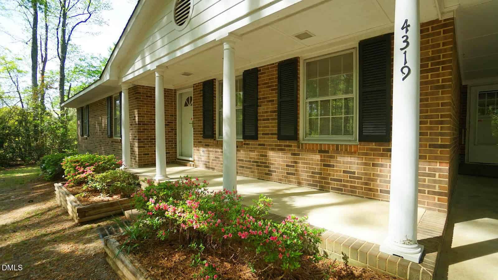 A brick house with black shutters, white columns, and a covered front porch. Pink flowers and shrubs line the walkway. The address 4319 is on a column near the carport.