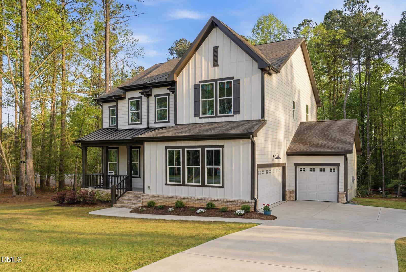 Two-story modern farmhouse with white vertical siding, black trim, covered front porch, attached two-car garage, and a neatly landscaped front yard, surrounded by tall trees.