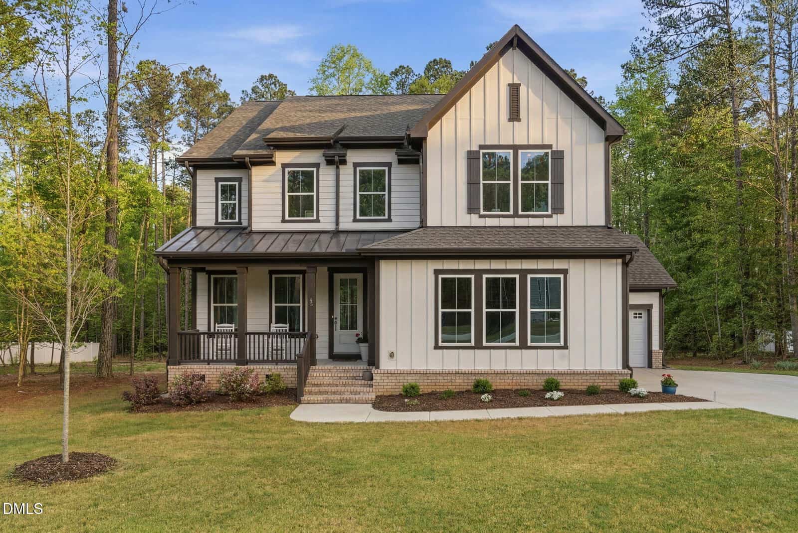 A modern two-story house with white vertical siding, dark trim, a covered front porch, and a manicured lawn. The home is surrounded by tall trees and has a concrete driveway on the right side.