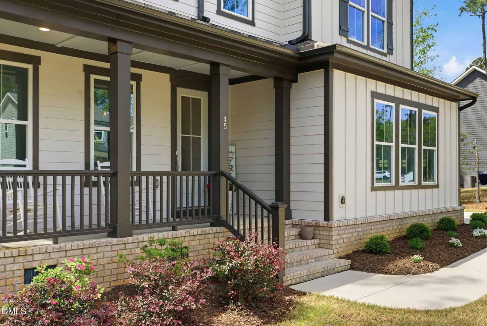 A modern house with light-colored siding and brick foundation, featuring a dark-trimmed front porch with railings, steps leading up, landscaped shrubs, and large windows. A concrete walkway curves toward the entrance.