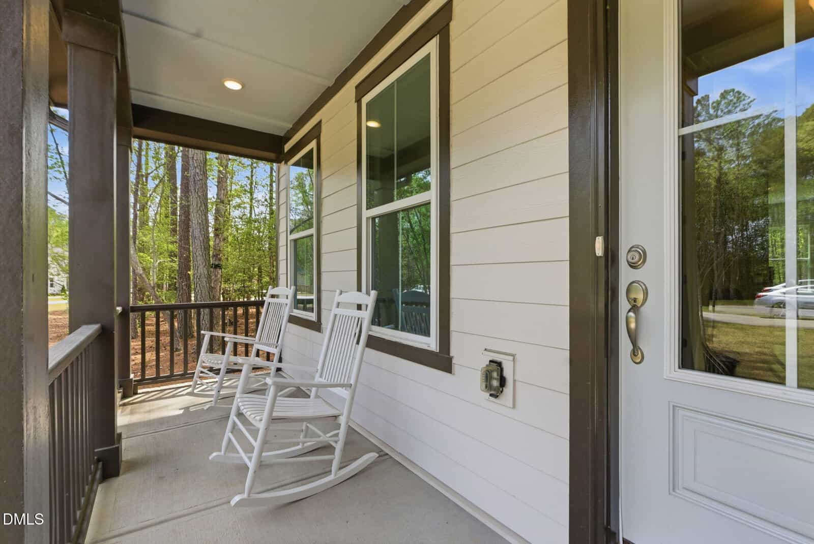 A cozy front porch with two white rocking chairs, brown railing, and beige siding, overlooking a wooded area with tall trees. The porch has a glass-paneled door and large windows.