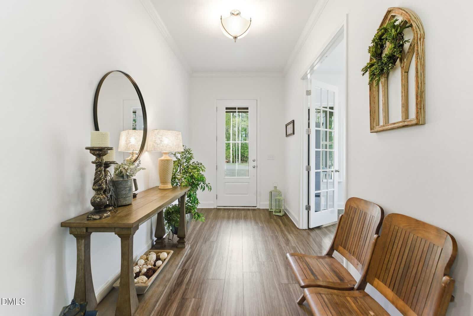 A bright hallway with wood flooring, a console table with decorative items and a mirror on the left, two wooden chairs on the right, and a white door with glass panels at the end.