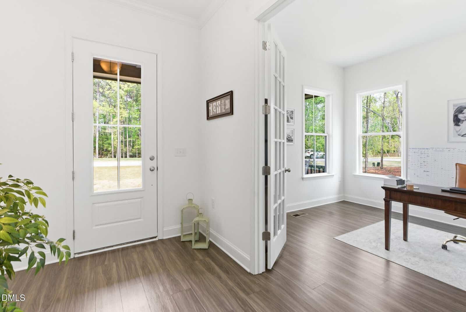 Bright, modern home office with wood floors, white walls, a large desk, windows letting in natural light, and glass French doors. A potted plant, lanterns, and wall art decorate the space.