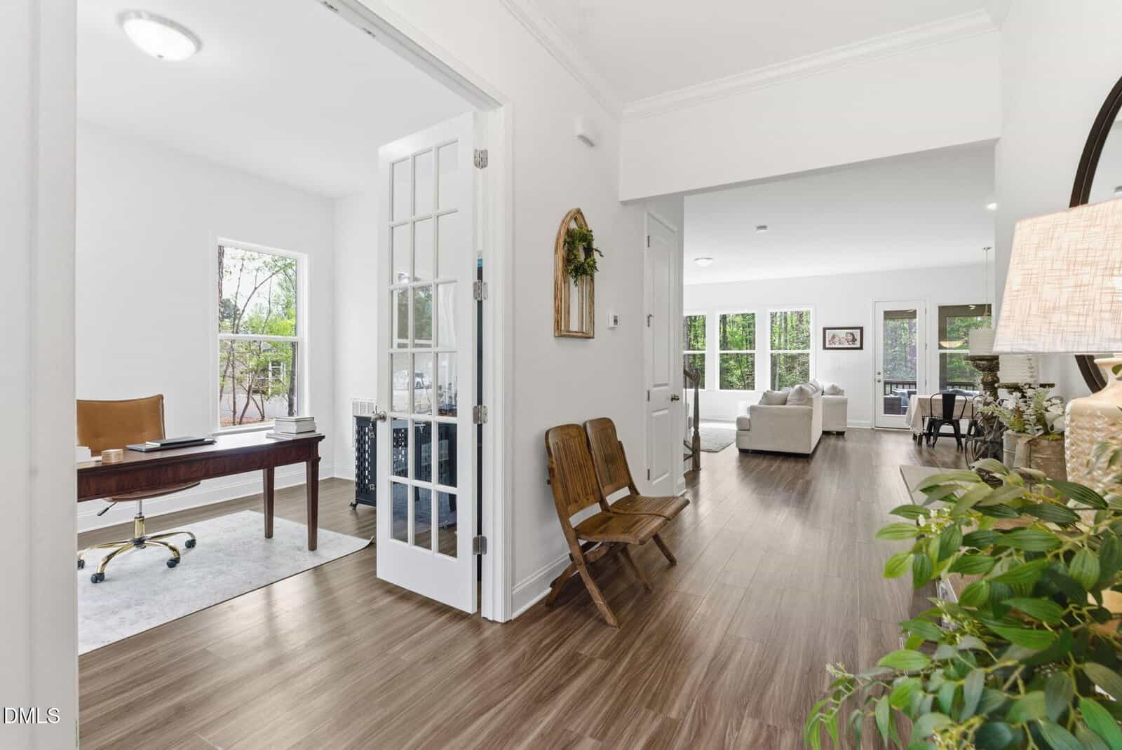 Bright entryway with wood floors, two wooden chairs, plants, and a round mirror. To the left, a home office with a desk and window; ahead, an open living room with large windows and light-colored furniture.