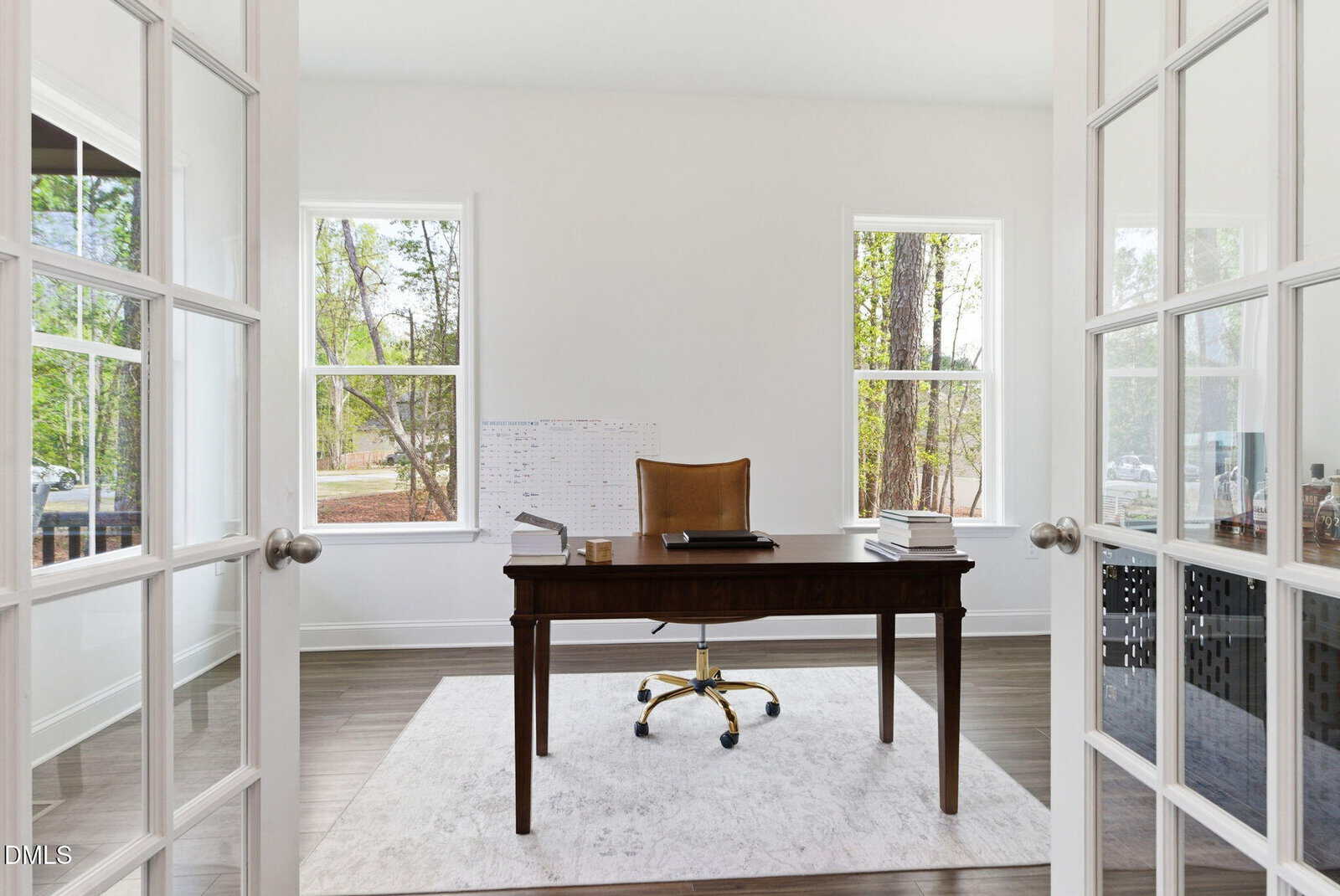 A modern home office with a wooden desk, brown chair, and two windows bringing in natural light. The desk has stacks of papers, a notebook, and a calendar. The room has light walls and double glass doors.