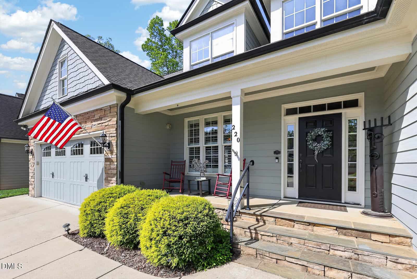 A modern suburban house with light blue siding, a black front door, and a porch featuring rocking chairs and a wreath. An American flag hangs near the garage, and manicured bushes line the front walkway.
