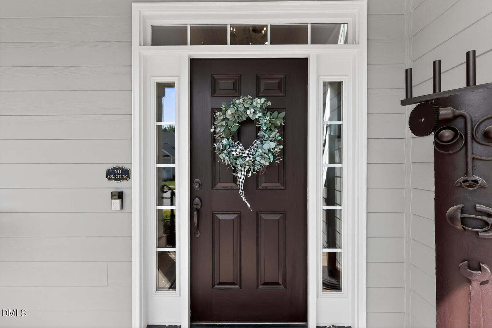 A dark brown front door with a wreath of green foliage, surrounded by white trim and glass panels; a "No Soliciting" sign is on the left, next to a doorbell, and a metal sculpture is on the right.