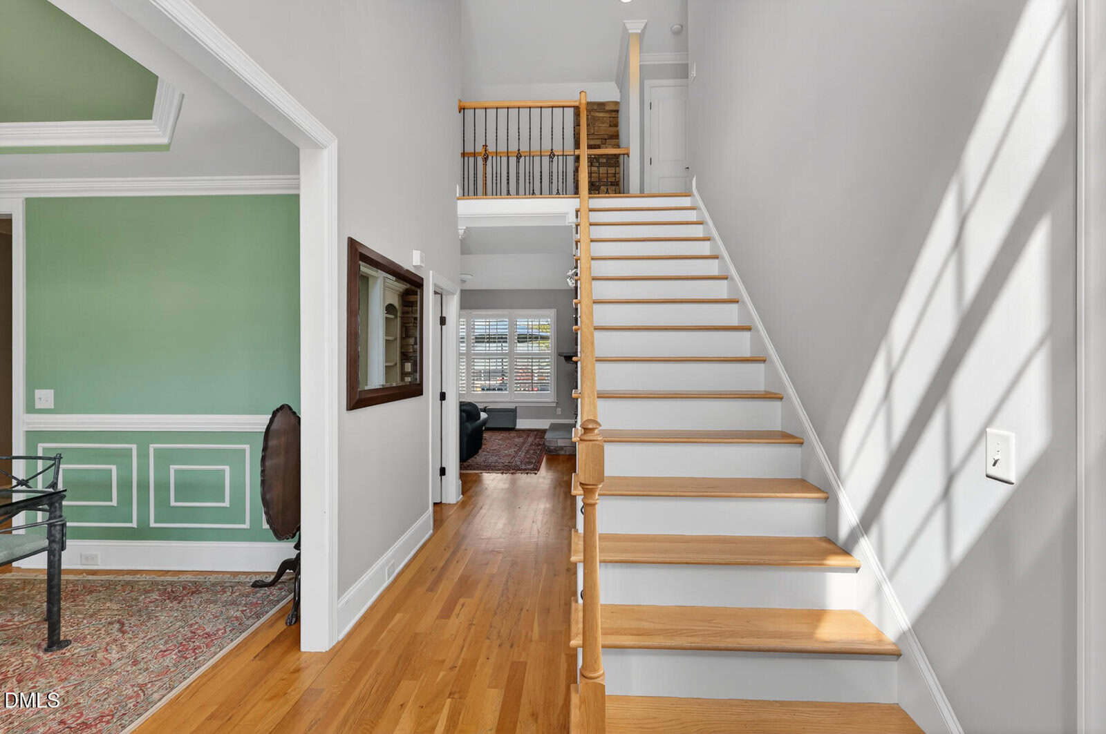 A bright hallway with wooden floors features a staircase with white risers and a wooden handrail, leading to an upper level. To the left, a green room with white trim and a large window is partially visible. Sunlight streams in through windows.