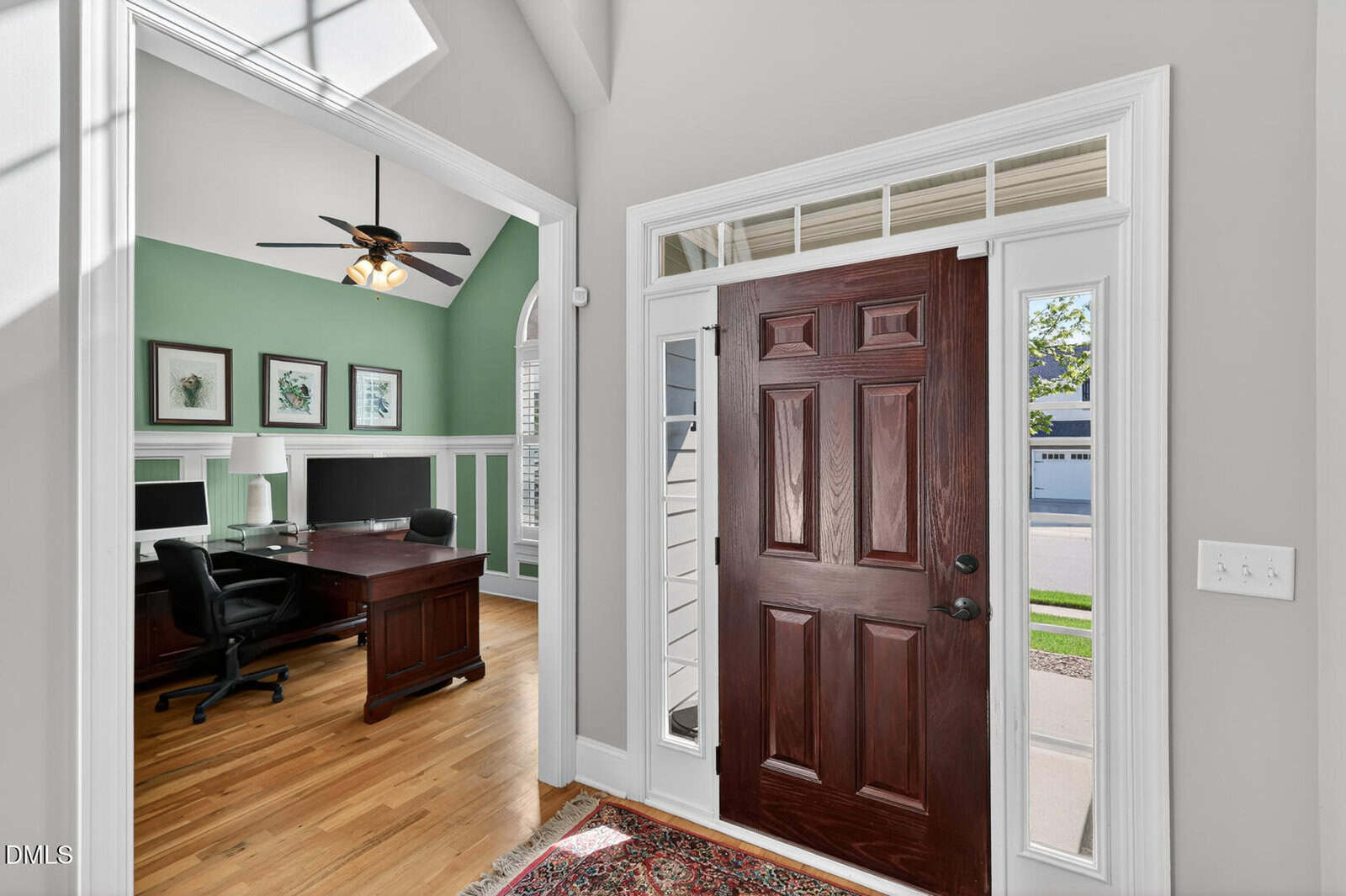 A home entryway with a dark wooden front door open, revealing a street outside. To the left is a home office with green walls, a wooden desk, and two computer monitors. The flooring is hardwood with a red rug near the door.