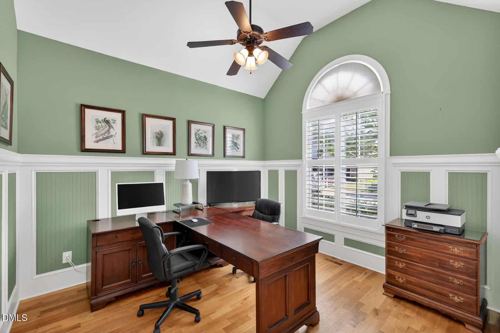A home office with green and white paneled walls, wooden desk, two monitors, black office chair, lamp, printer on a side cabinet, three framed art pieces, and a large arched window with plantation shutters.