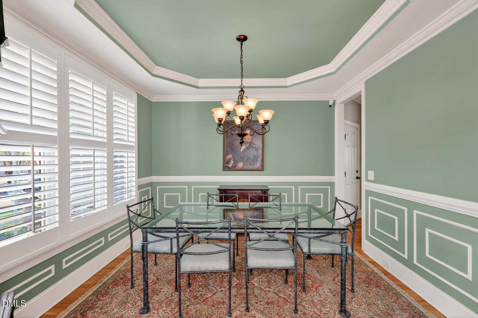A dining room with a glass table, six metal chairs, green walls with white trim, a chandelier, large windows with white shutters, and a framed picture above a small cabinet.