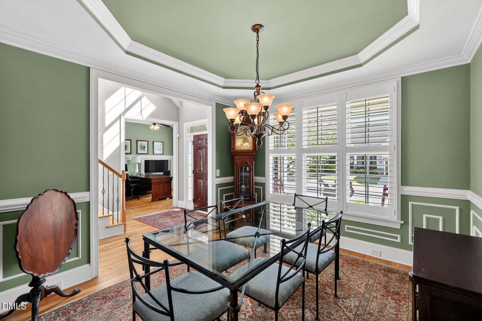 A dining room with green walls, white trim, a glass-top table with six chairs, a chandelier, and large windows with shutters. An open doorway leads to a foyer with a staircase and office area.