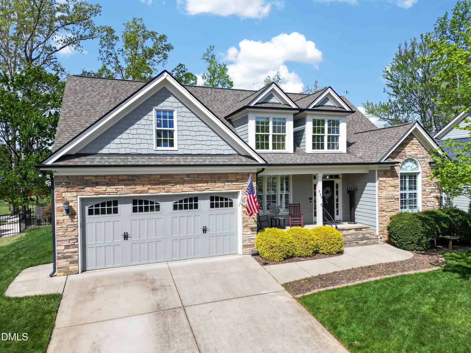 A two-story suburban house at 220 Plantation Drive in Youngsville, featuring gray siding, stone accents, a double garage, dormer windows, and a front porch with an American flag, surrounded by a green lawn and shrubs on a sunny day.