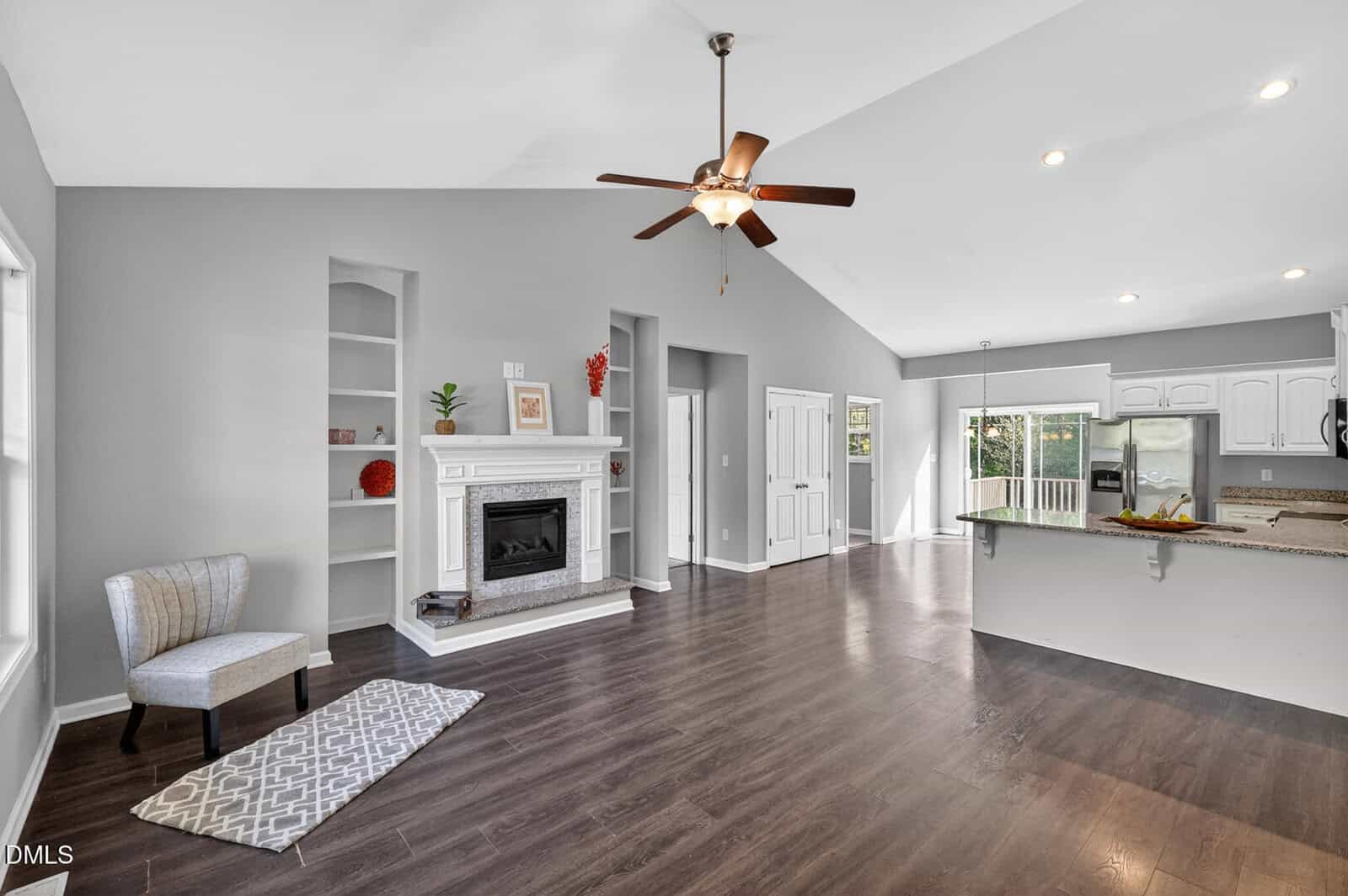 Open living space with dark hardwood floors, a white fireplace, built-in shelves, a chair and rug, ceiling fan, and a kitchen with white cabinets and a breakfast bar. Glass doors lead outside.
