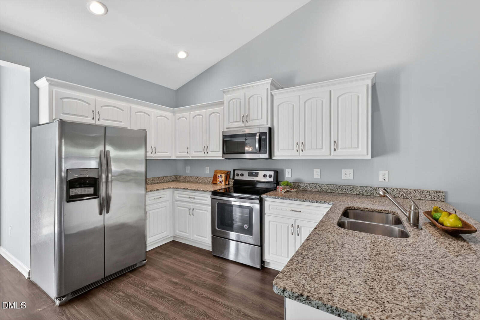 Modern kitchen with white cabinets, stainless steel appliances, granite countertops, double sink, and light gray walls. A fruit bowl and a book are on the counters. The floor is dark wood.