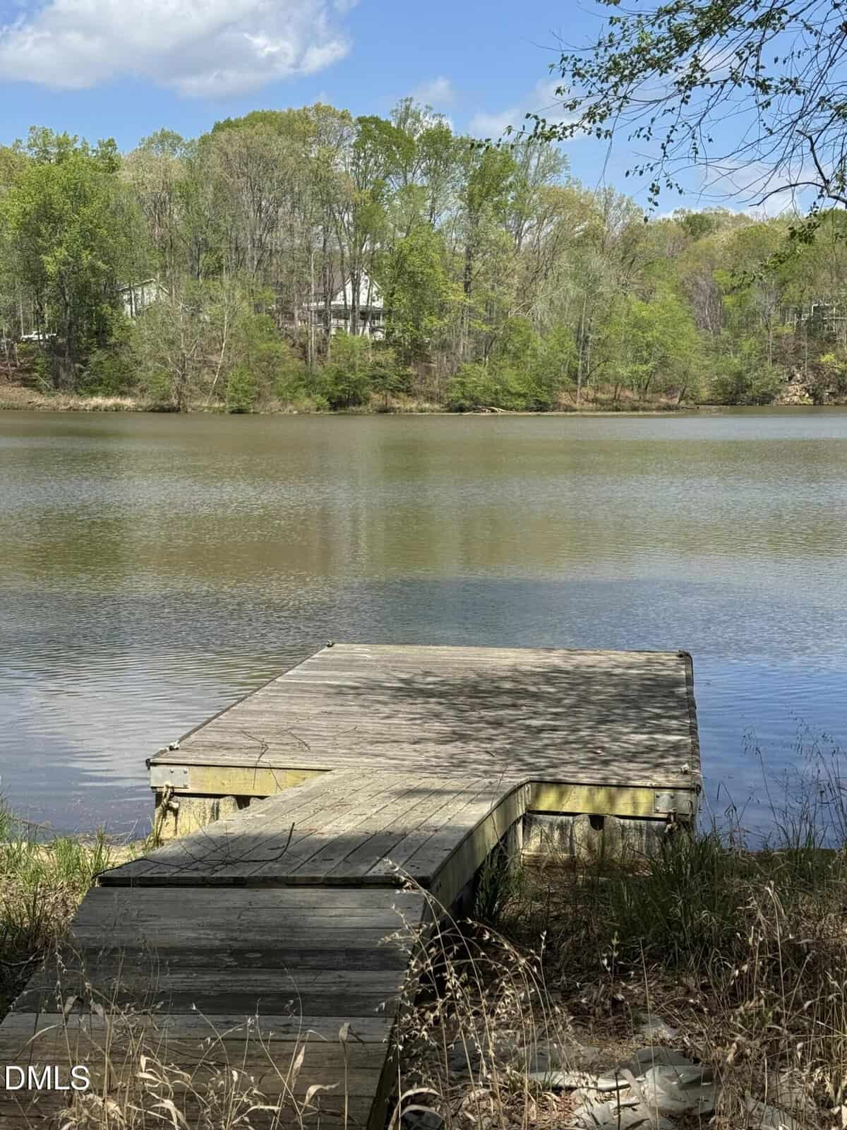 A small wooden dock extends into a calm lake surrounded by trees with green foliage near 00 Peninsula Drive in Henderson. A few houses are visible among the trees on the opposite shore under a partly cloudy sky.