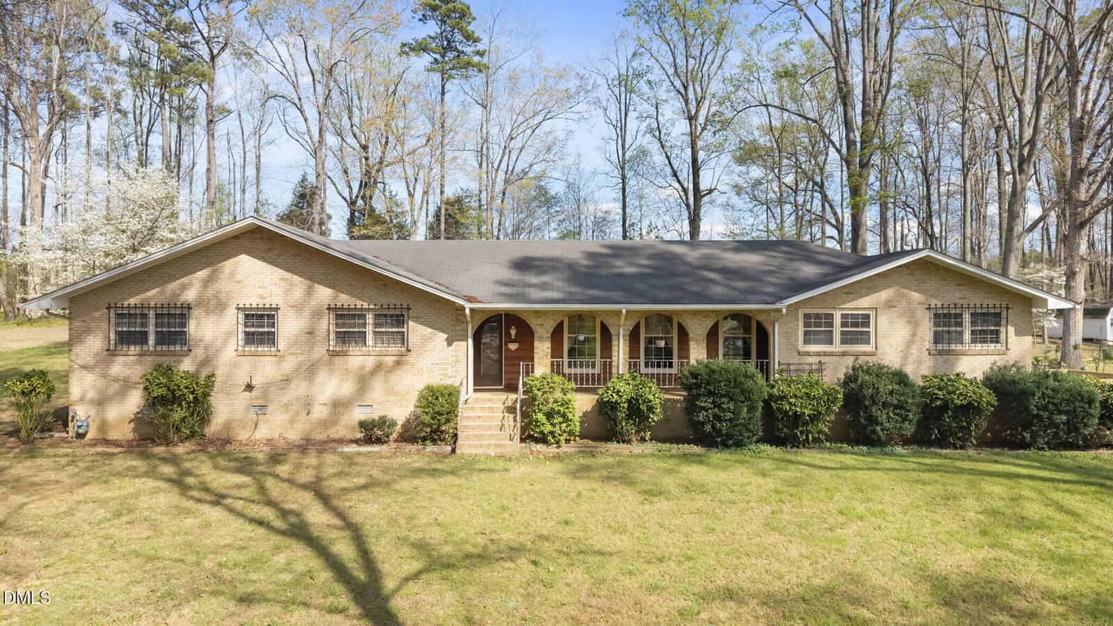 Single-story, light brick house with a dark roof, covered front porch with arches, and several windows, surrounded by bushes and trees, on a large grassy yard with woods in the background.