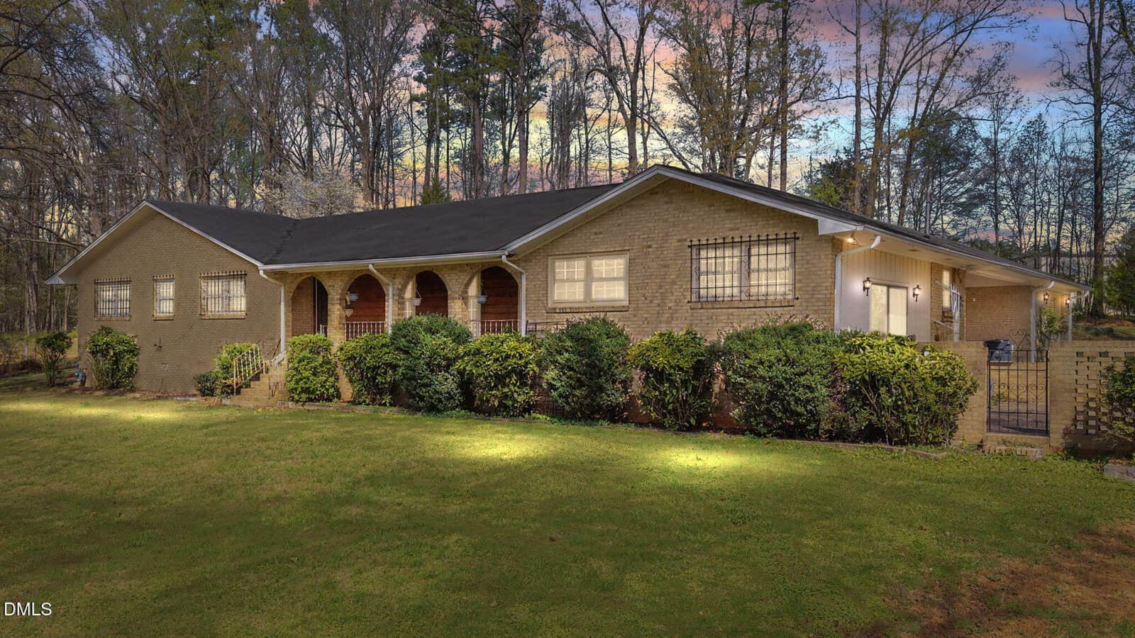 Single-story brick house with a black roof, arched front porch, and large windows, surrounded by bushes and grass. Trees and a colorful sunset sky are visible in the background.
