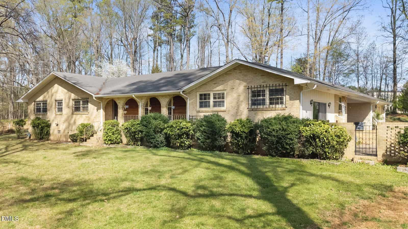 Single-story brick house with arched front porch, black railings, and multiple windows, surrounded by green bushes and a grassy lawn. Tall trees without leaves stand in the background.