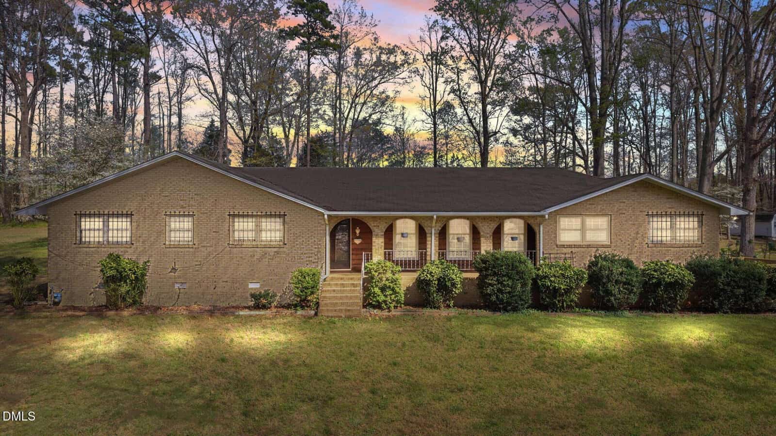A single-story brick house with a dark roof and arched front porch, surrounded by shrubs and trees, sits on a grassy lawn at sunset.