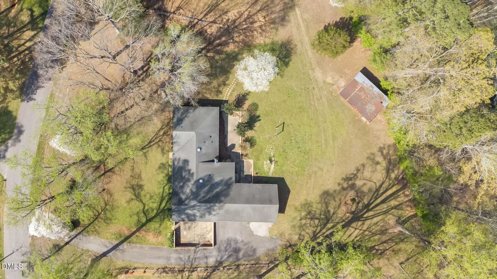 Aerial view of a house surrounded by trees with a paved driveway, a grassy yard, and a separate small, rusted-roof building nearby. Shadows from trees are cast across the property.