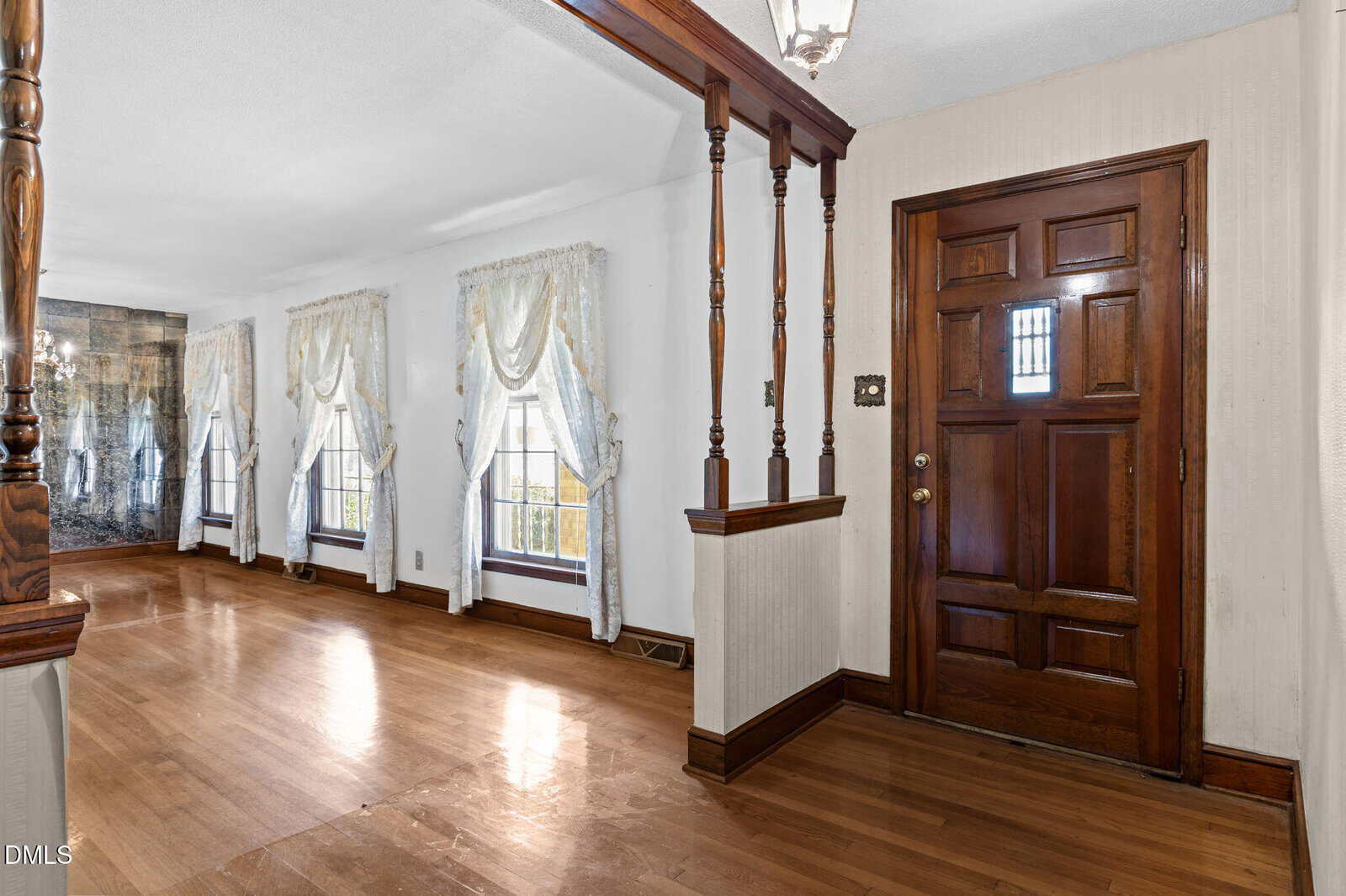 A wooden front door with a small window opens to a hallway with hardwood floors, three tall windows with white curtains, and decorative wooden columns separating the entry from the living area.