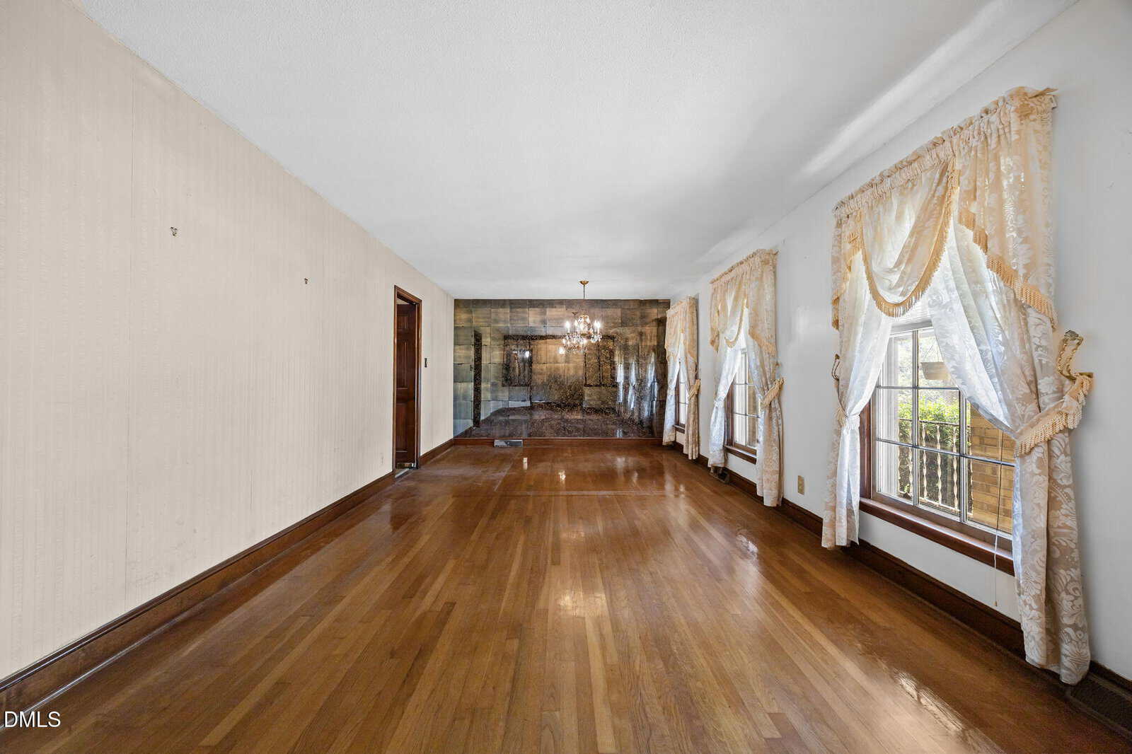 Empty room with hardwood floors, cream-colored walls, two large windows with sheer curtains on the right, a wooden door on the left, and a chandelier in the far area near a mirrored wall.