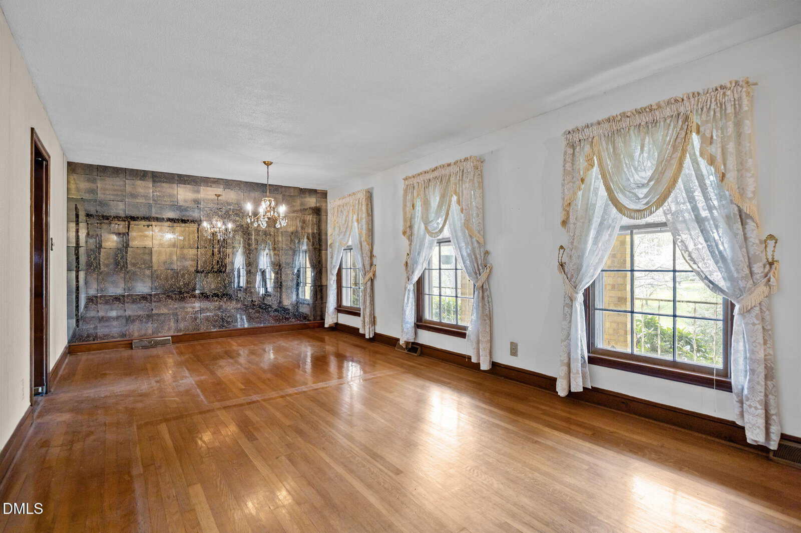 Empty room with polished wood floors, two large windows with sheer lace curtains, and a mirrored wall featuring a chandelier. Walls are white, and natural light enters from the windows on the right side.