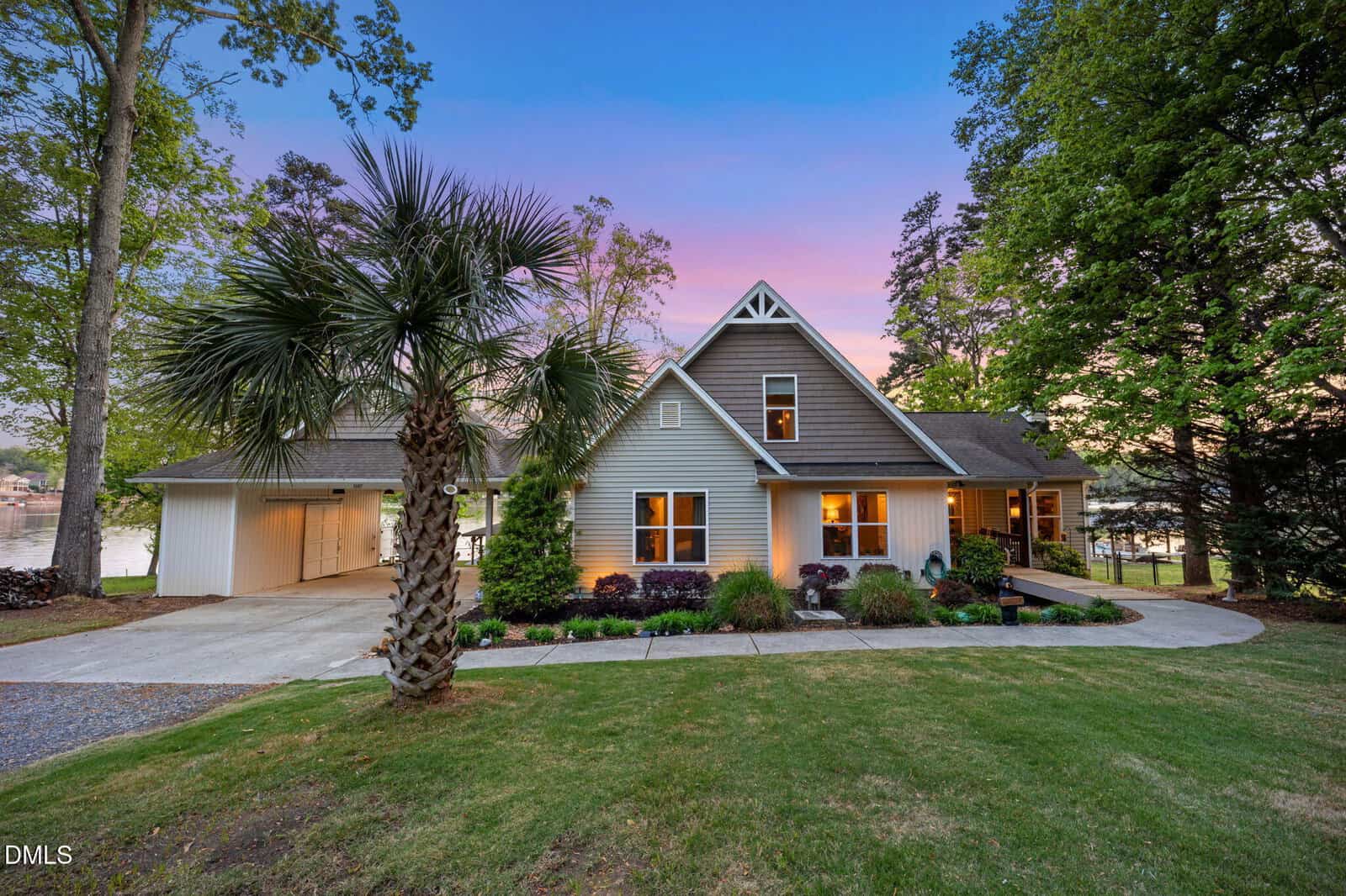A two-story house with beige siding and a covered carport sits on a green lawn at 1187 Sagamore Drive in Louisburg, surrounded by tall trees. A palm tree stands in the foreground as the sky highlights a pink and blue sunset.