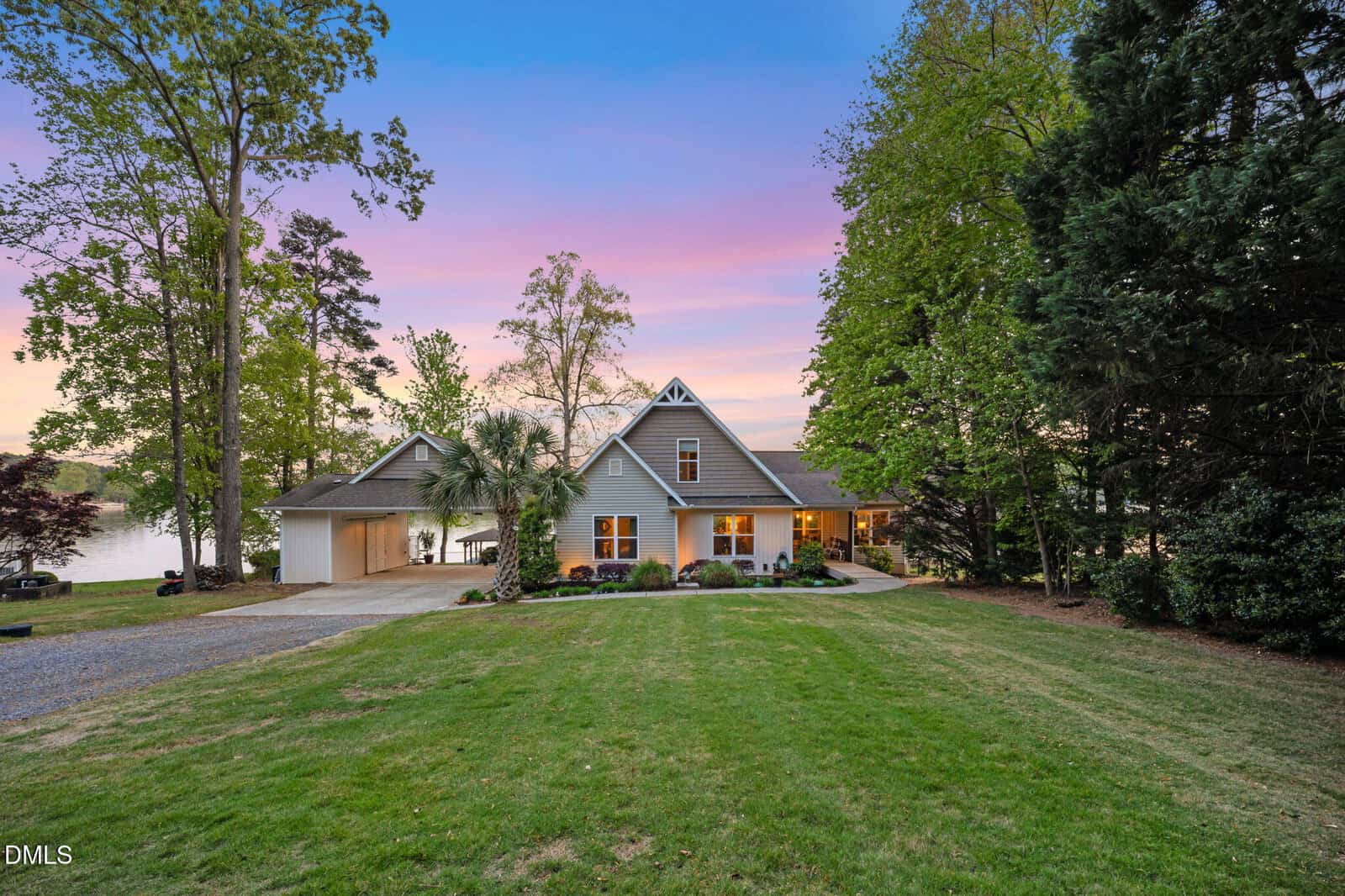 A gray house with a gabled roof and attached garage sits among trees at sunset. The front yard is grassy, and a lake is visible in the background on the left side.