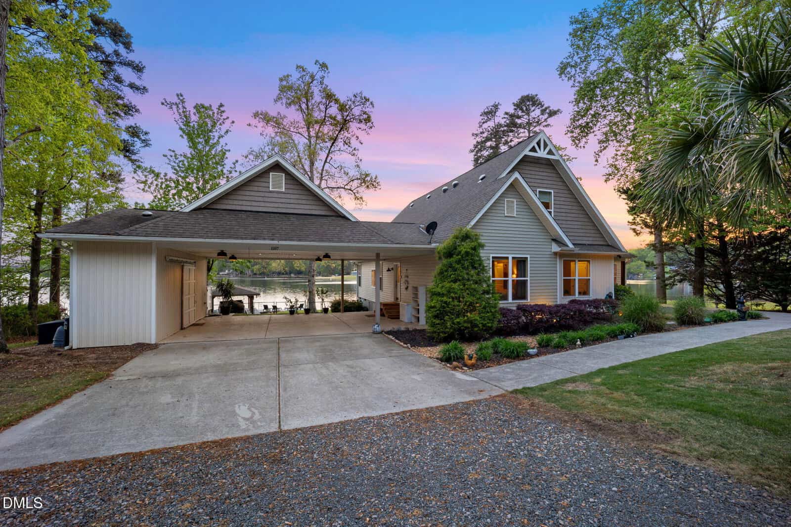 A gray house with dormer windows and a double carport sits beside a lake, surrounded by tall trees and a manicured lawn. The sky is purple and pink at sunset. A paved driveway leads to the entrance.