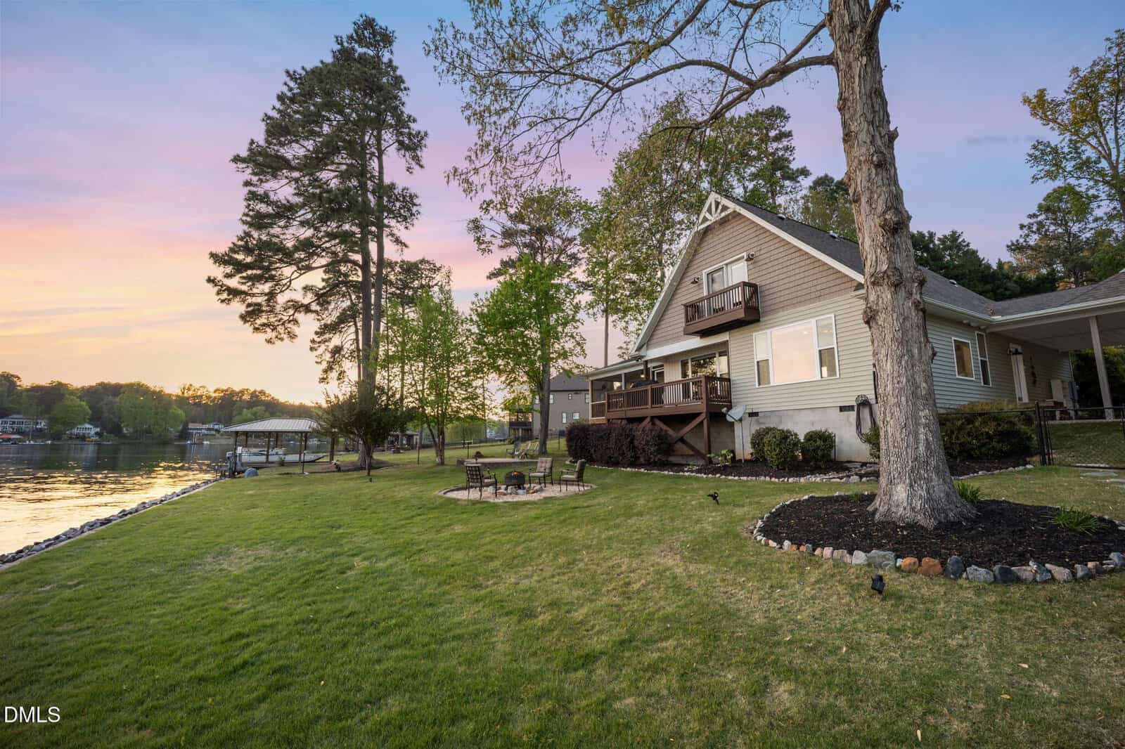 A lakeside house with a wooden deck, surrounded by trees and grass, sits near the water at sunset. A fire pit with chairs is placed on the lawn, and a boat dock can be seen in the distance.